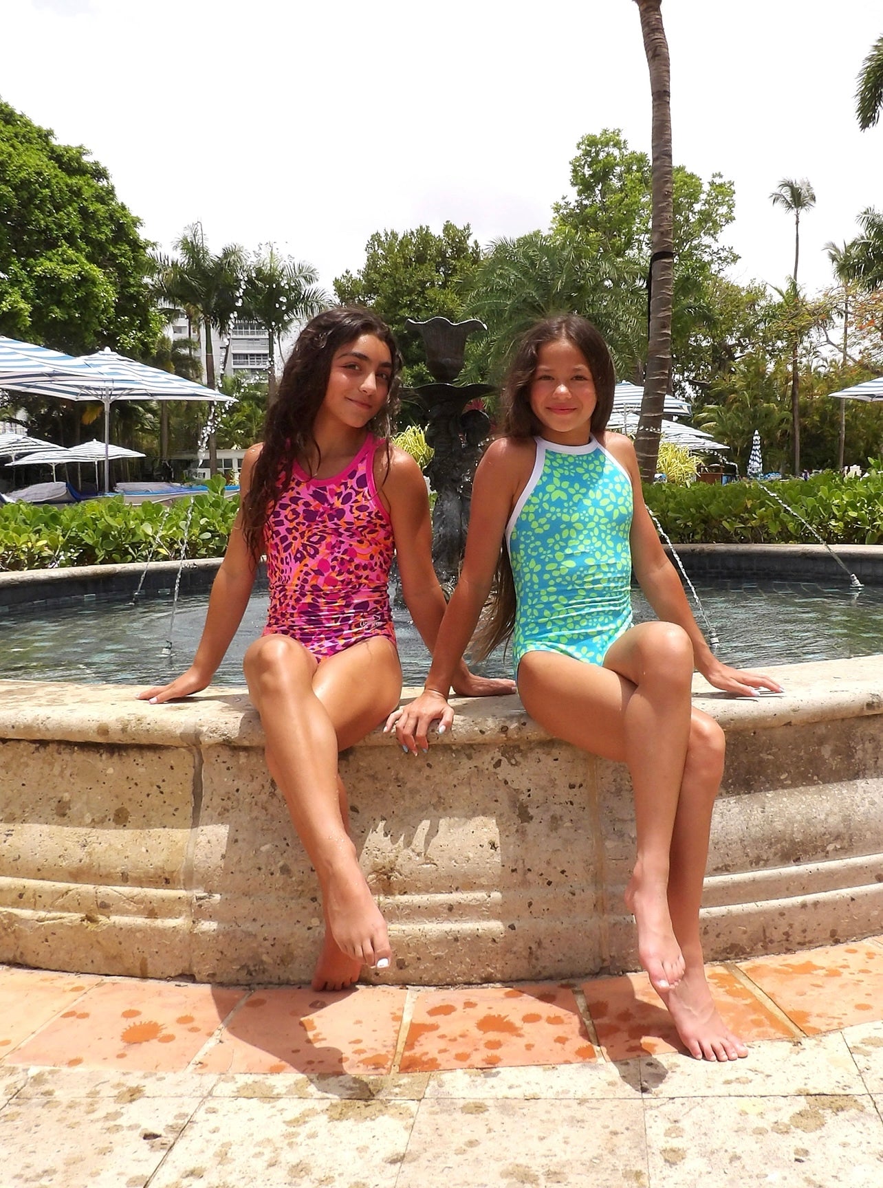 Two  girls in leotards sitting on a stone ledge by a pool with trees and umbrellas in the background.