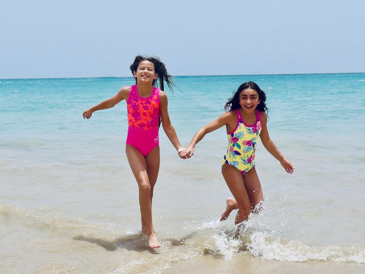 Two girls in colorful leotards running on a beach with clear blue water and sky. wearing Mariposas and Kalani Floral leotards