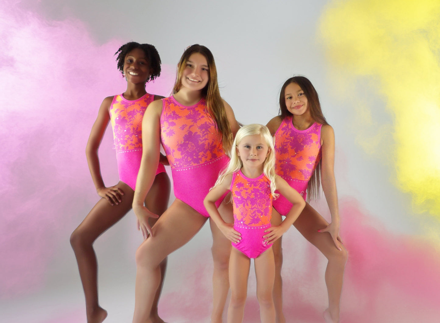 Four girl wearing pink and orange leotards against a white background with colorful smoke effects.