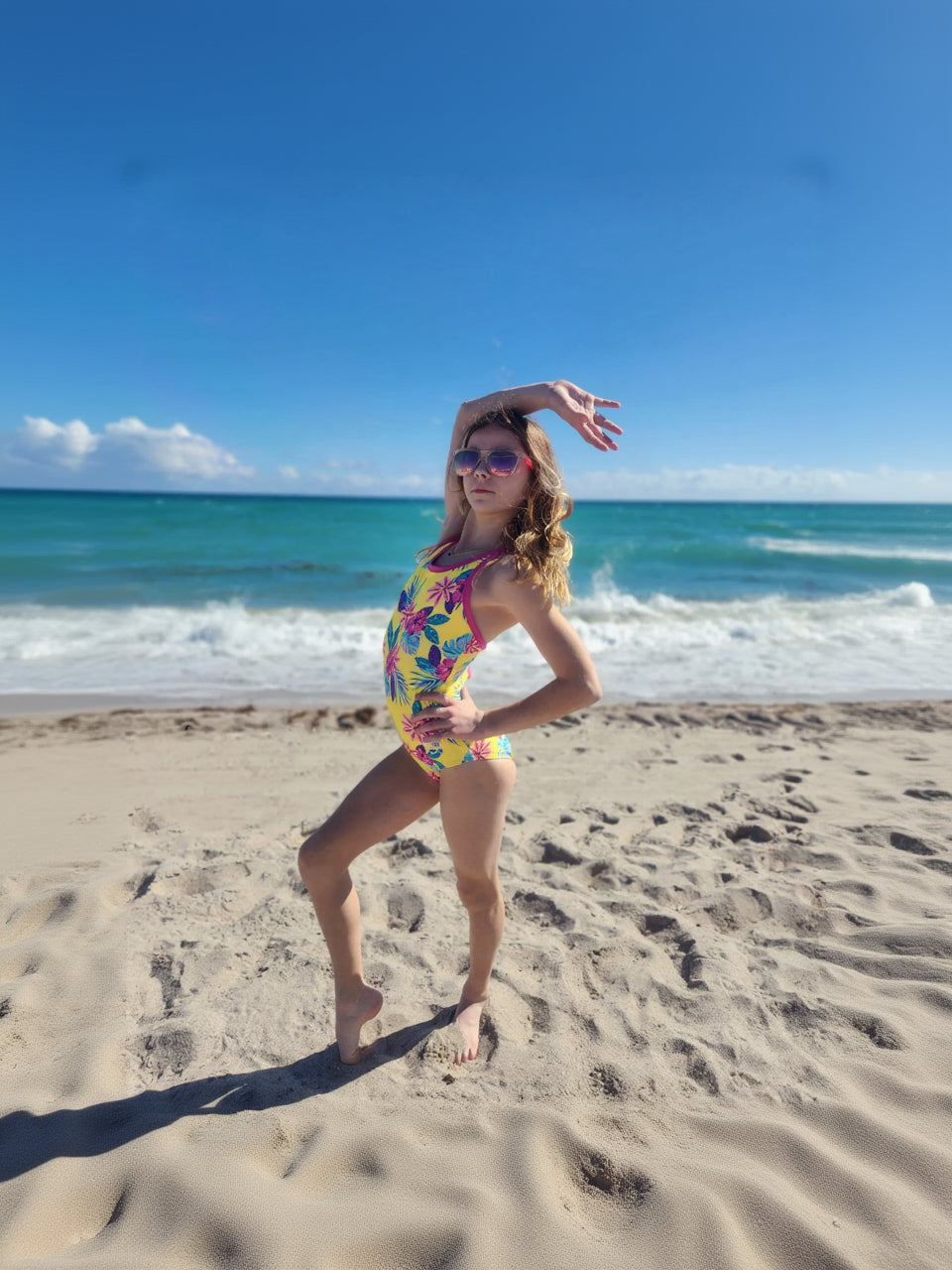 Girl in a colorful leotard posing on a beach with blue sky and ocean.
